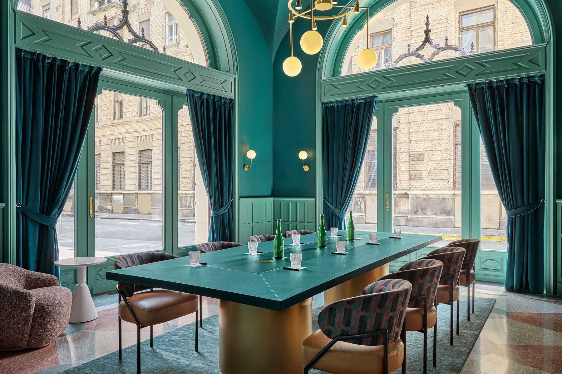 Chic, modern room with teal walls and large arched windows, a long teal table with glasses and water bottles, patterned chairs, globe pendant lights, and dark teal curtains. Bright natural light fills the space.
