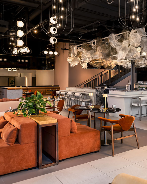 A modern restaurant interior with orange couches, wooden chairs, a white marble bar, and artistic hanging light fixtures. Green plants add a touch of nature and large, airy sculptures hang from the ceiling.
