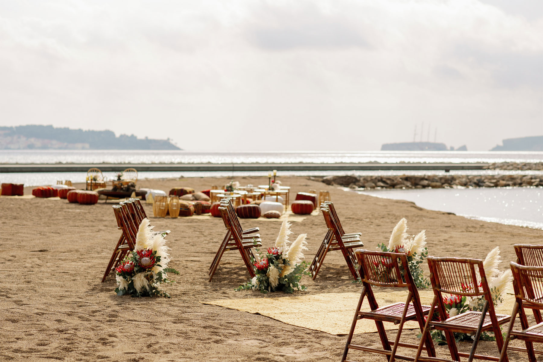 Wooden chairs decorated with flowers are arranged on a sandy beach, facing the sea. Low tables and cushions are set up further back, with calm water and distant ships visible under a cloudy sky.