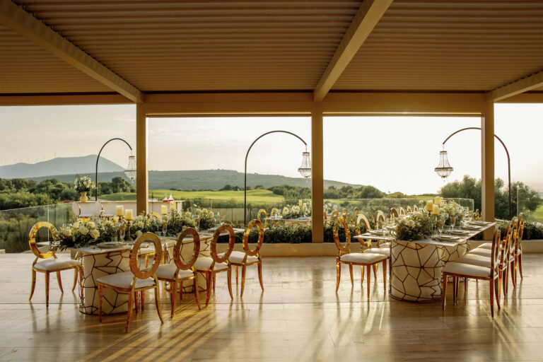 Elegant outdoor dining area with gold and white chairs around floral-decorated tables, overlooking a green landscape and hills, under a covered patio with large open windows and hanging chandeliers.