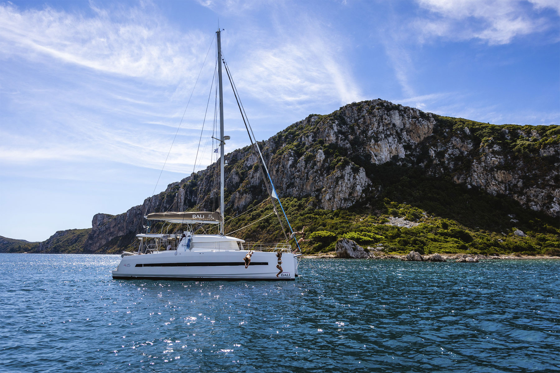 A white sailboat floats on calm blue water near a rocky, green hillside under a partly cloudy sky, with a person climbing from the water onto the boat.