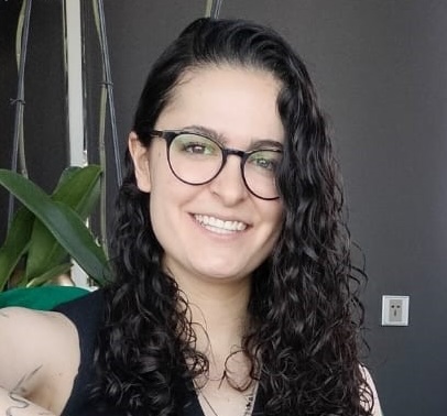 Una persona con el pelo largo, rizado y oscuro y gafas sonríe a la cámara. Lleva un top negro sin mangas y está de pie en un interior con una planta y una pared oscura de fondo.