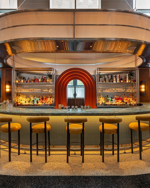 A modern bar with a curved counter, yellow barstools, and illuminated shelves stocked with bottles. The bar features an arched orange accent and contemporary lighting, creating a stylish, inviting atmosphere.