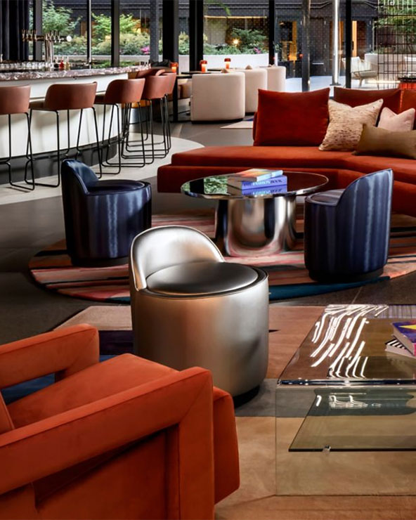 A modern lounge with colorful furniture, including orange chairs, a metallic silver chair, round tables, and a curved sofa. The space features floor-to-ceiling windows and a bar area with high stools in the background.