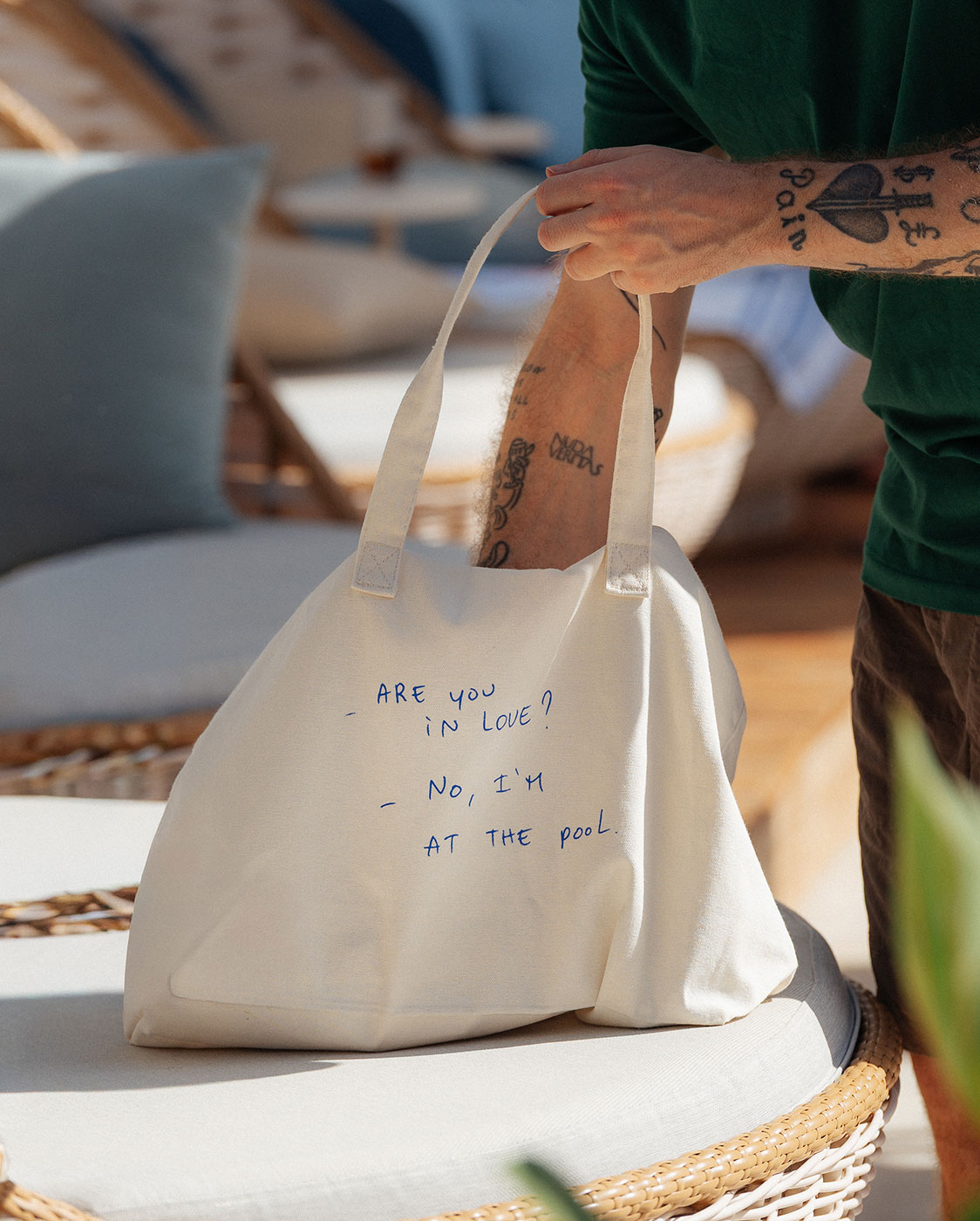 A person with tattooed arms, wearing a green shirt, places a white canvas bag with blue handwritten text reading 