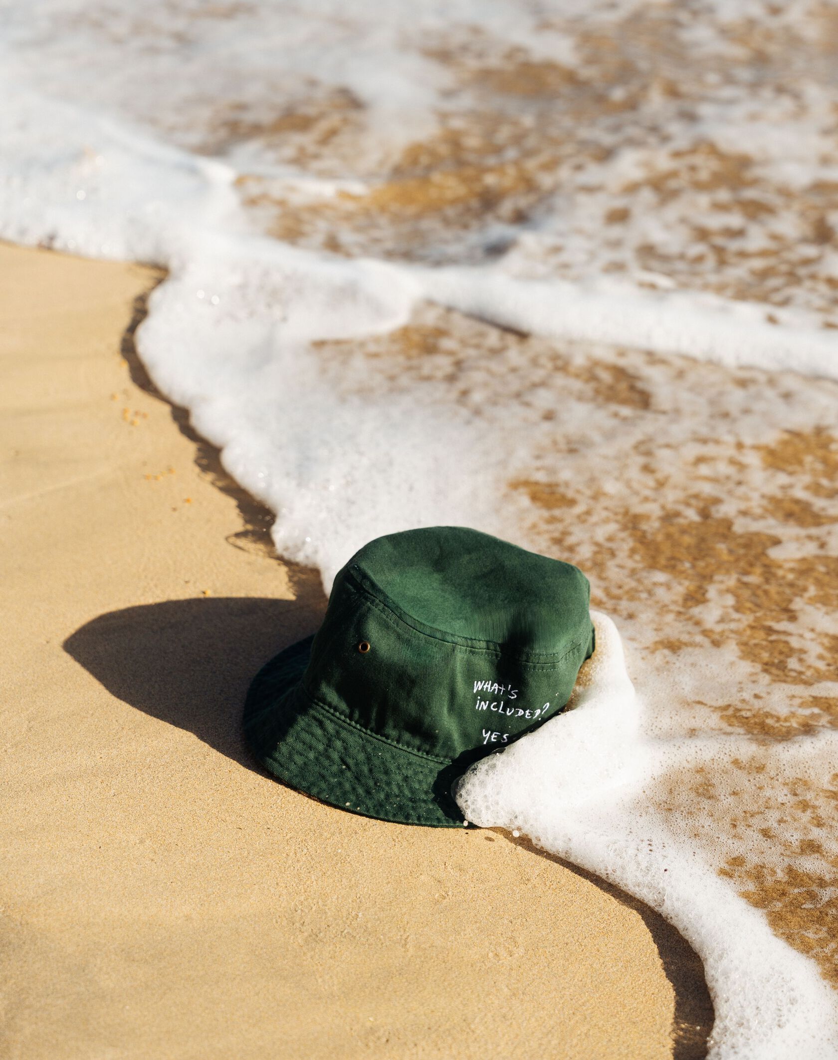 A green bucket hat partially buried in wet sand as ocean waves gently wash over it on a sunny beach.