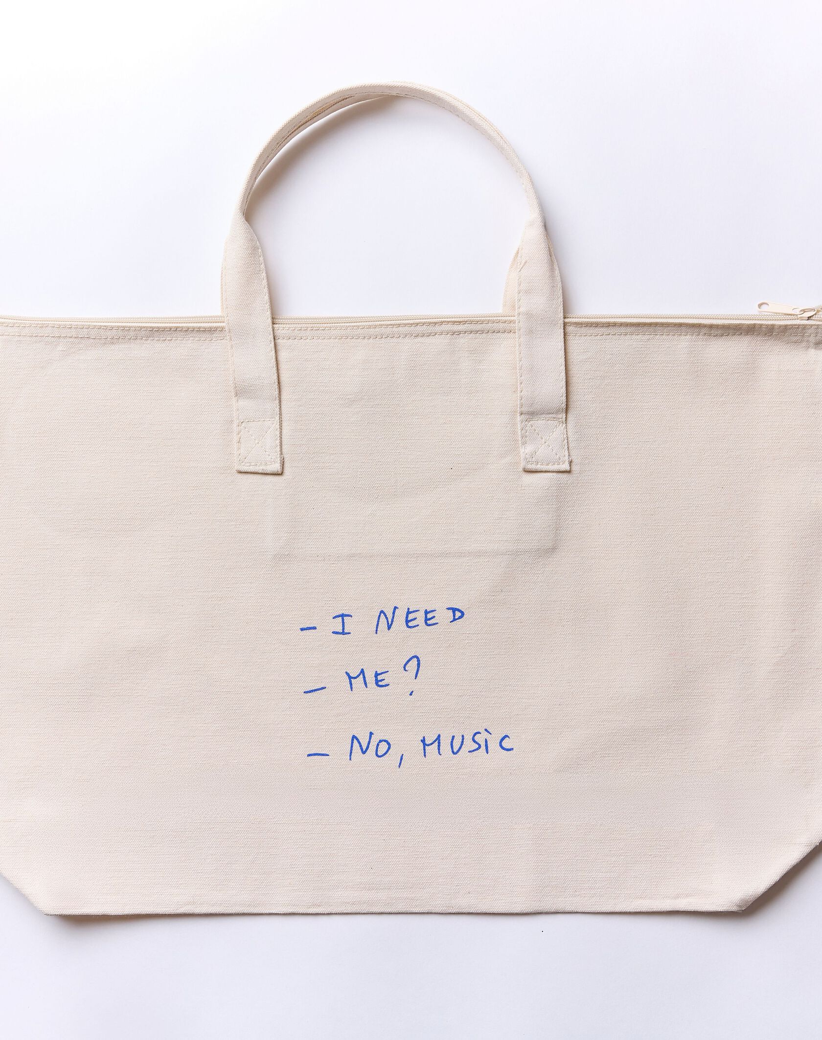 A beige canvas tote bag with blue handwriting that reads: – I NEED – ME? – NO, MUSIC.