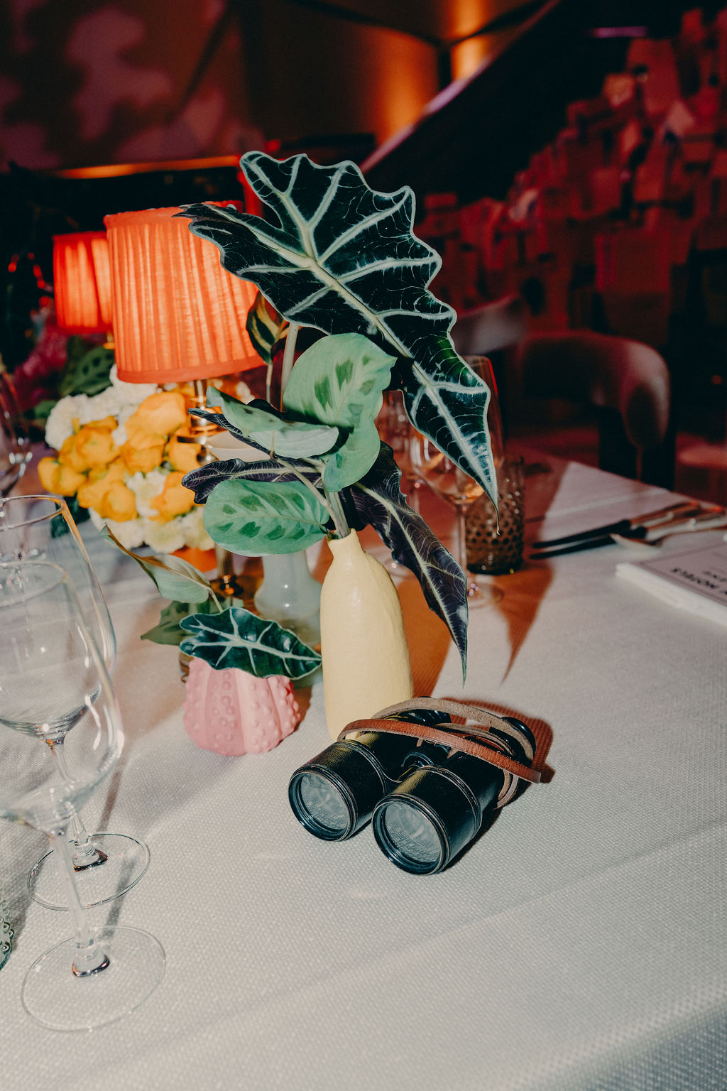 A pair of black binoculars with a brown strap sit on a white tablecloth next to a vase with green leafy plants, a pink vase with yellow flowers, orange lamps, and glassware.