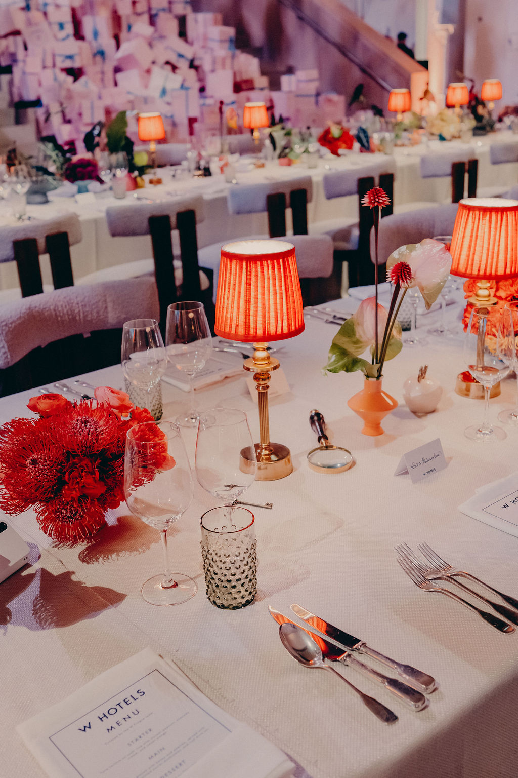Elegant table setting at a formal event, featuring red lamps, vibrant red and pink flower arrangements, glassware, menus, and name cards on white tablecloths. Rows of similar tables appear in the background.