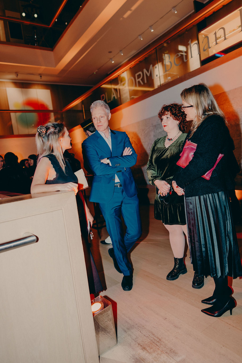 Four adults dressed in formal attire stand and converse in a modern, warmly lit indoor setting with art on the walls. One man in a blue suit stands with three women, two in dark dresses and one in green velvet.