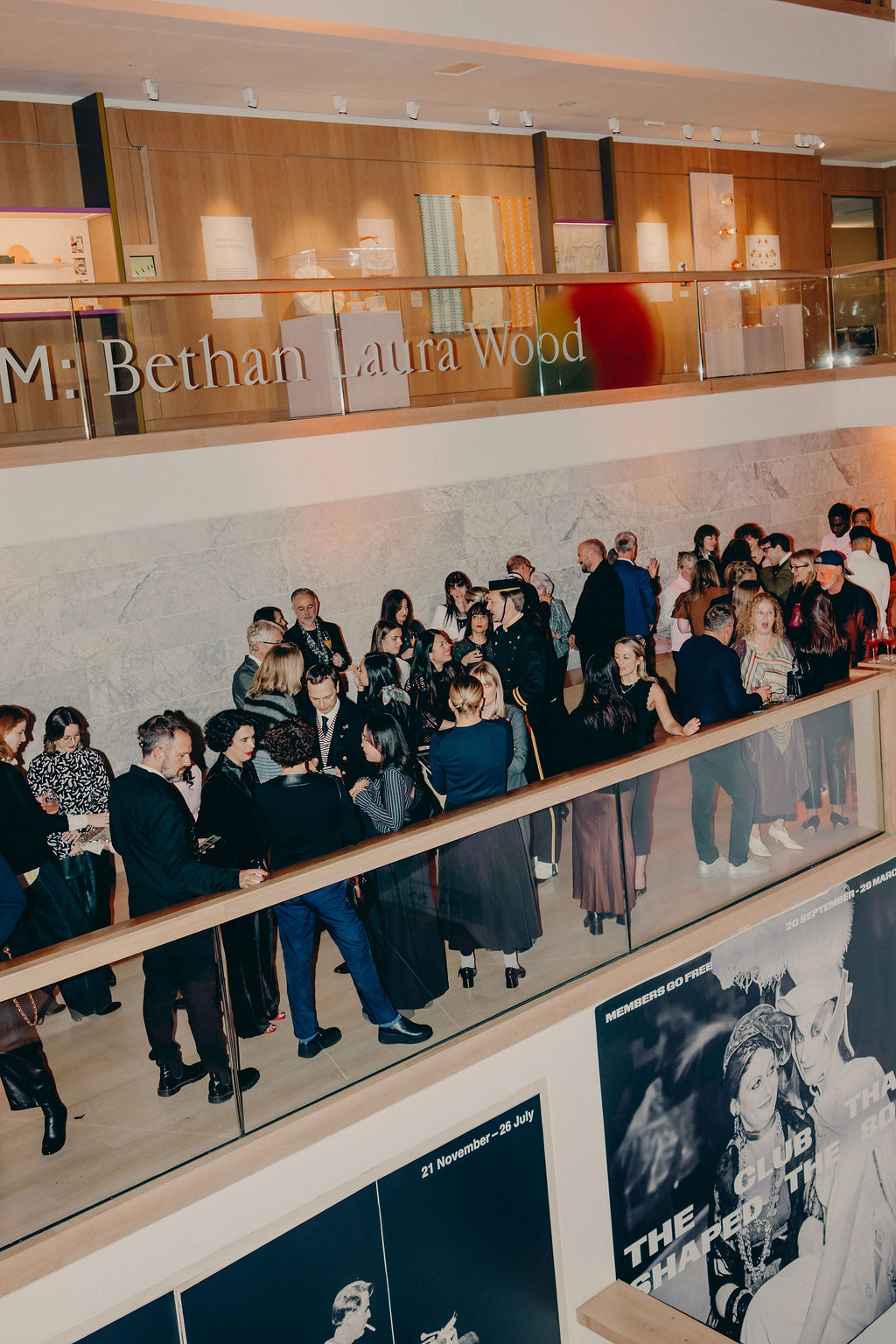 A crowd of people in formal attire mingles at an indoor event in a modern building with posters and signage for Bethan Laura Wood visible on the upper level.