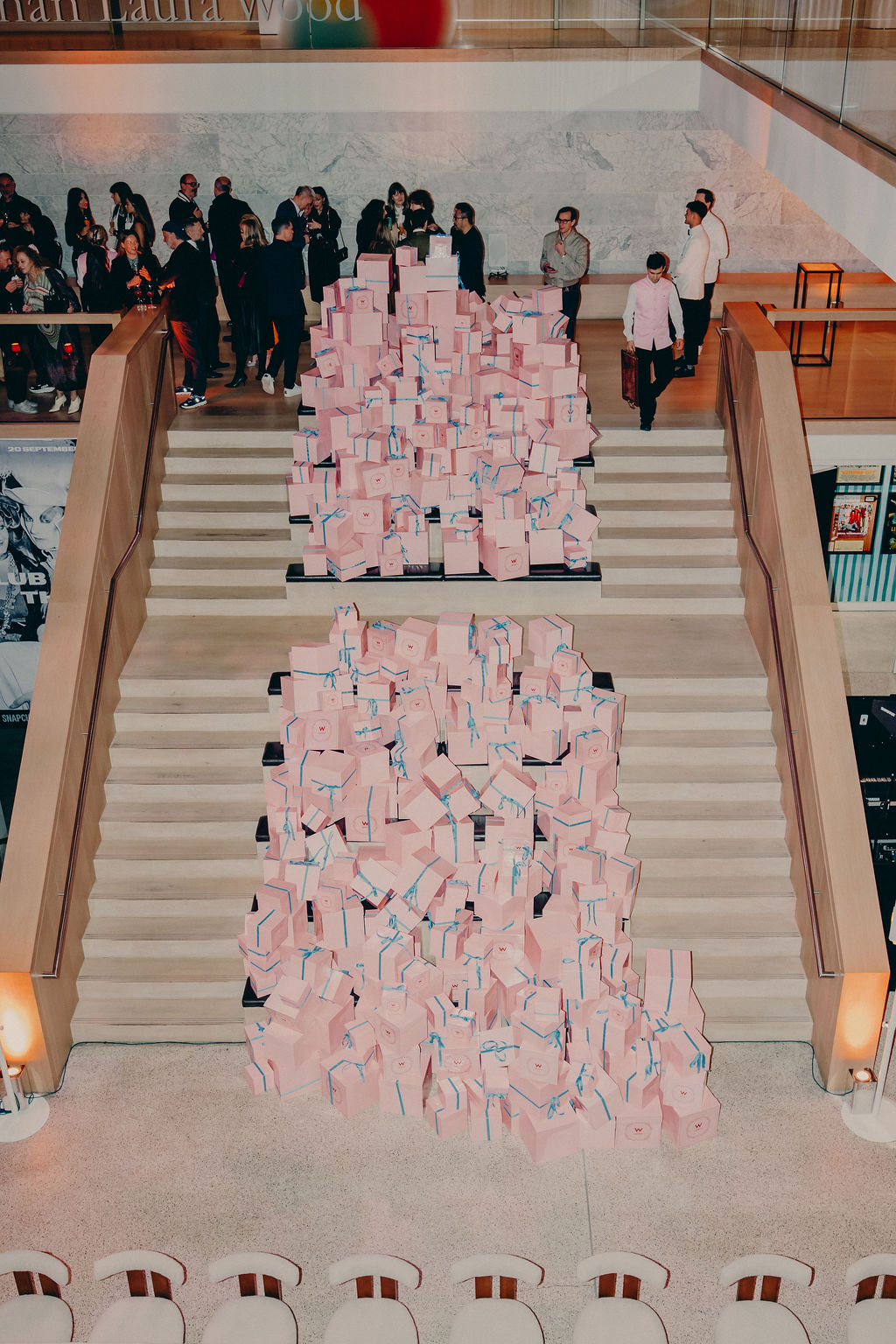 Large staircase covered with stacked pink boxes, some tied with blue ribbon, leading up to a landing where a crowd of people are gathered. The setting appears to be an indoor public space or gallery.