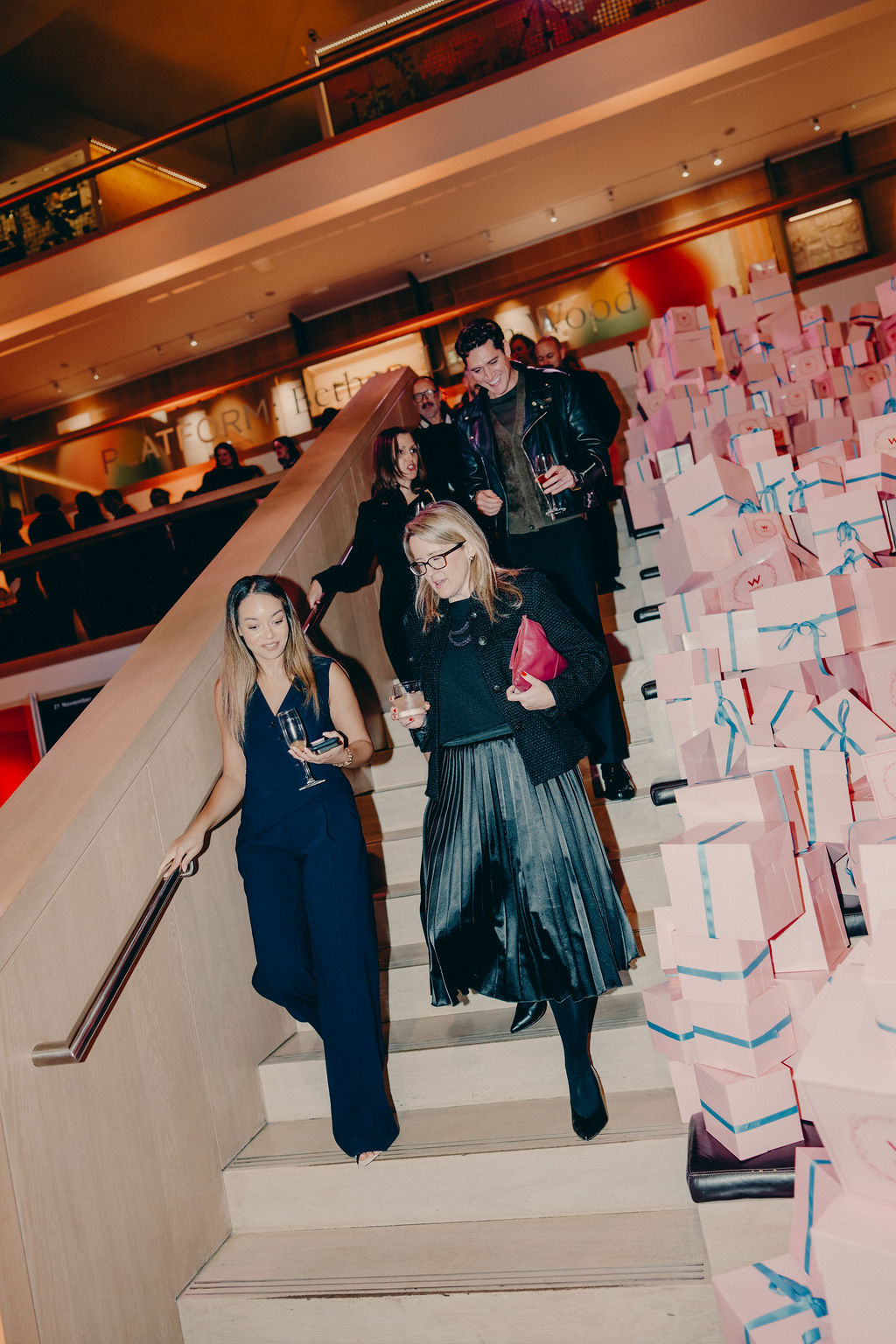 A group of people dressed formally descends a staircase, holding drinks. The stairwell is decorated with stacks of pink gift boxes tied with blue ribbons. The setting appears to be an upscale indoor event or party.
