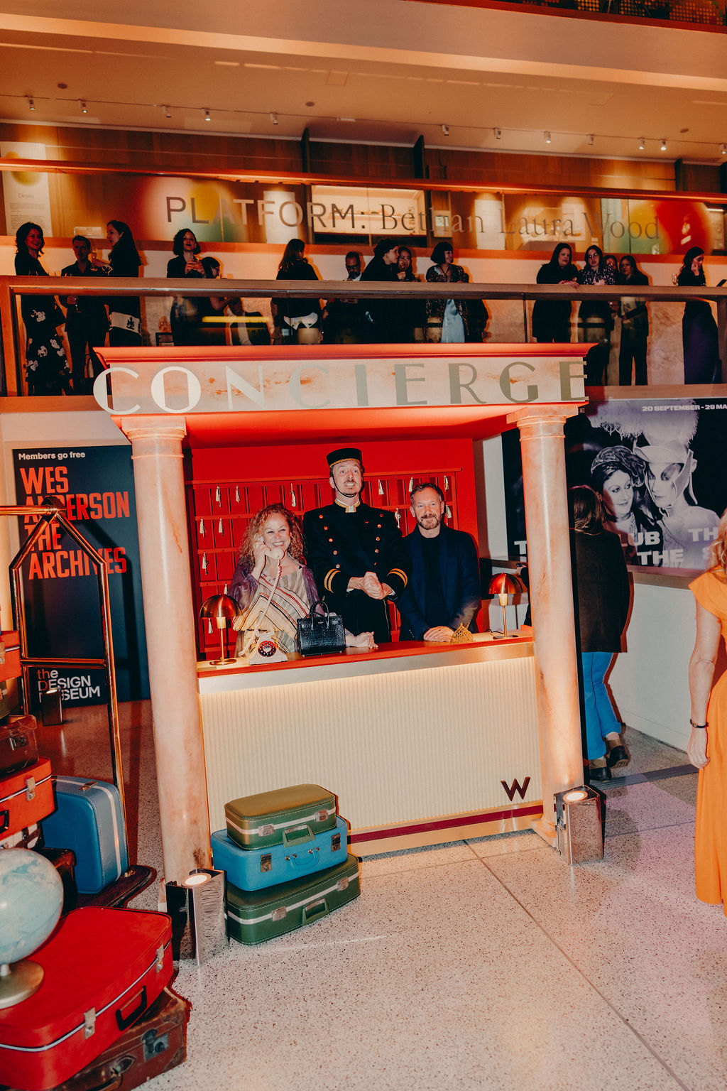 Three people stand behind a themed concierge desk with suitcases stacked in front. They are indoors at an event, with colorful lights and posters in the background, and people visible around them.