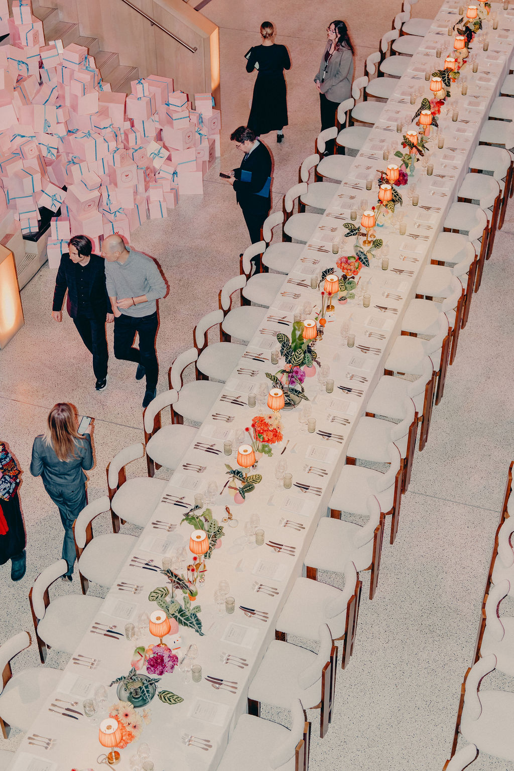 A long, elegant dining table is set with floral centerpieces and candles, surrounded by empty chairs. Several people stand nearby, and pastel-colored gift boxes are stacked in the background. The scene appears to be at an event venue.