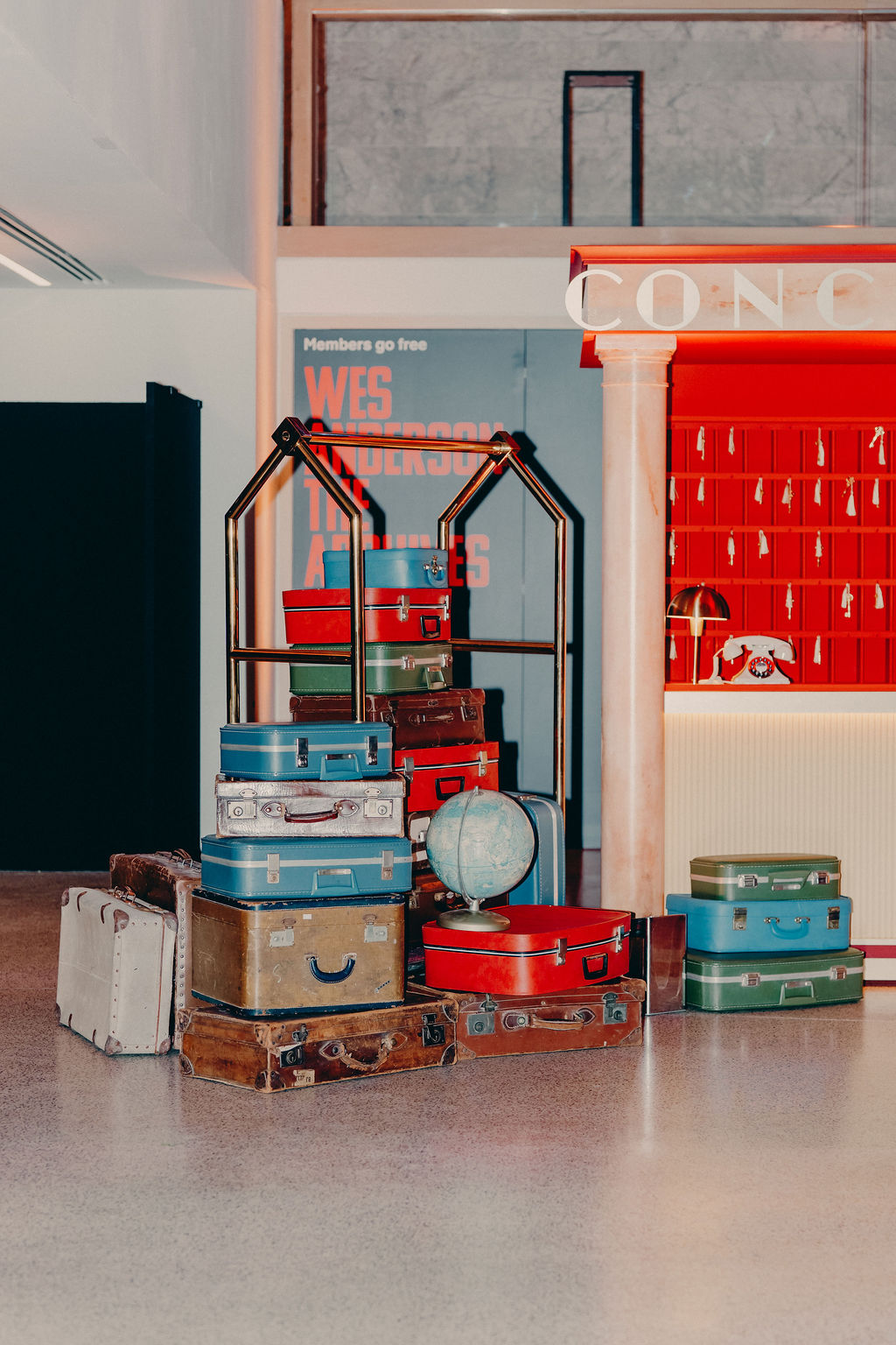 A colorful pile of vintage suitcases and a globe sit on a polished floor near a red hotel reception desk and a display sign in a stylish, modern interior.