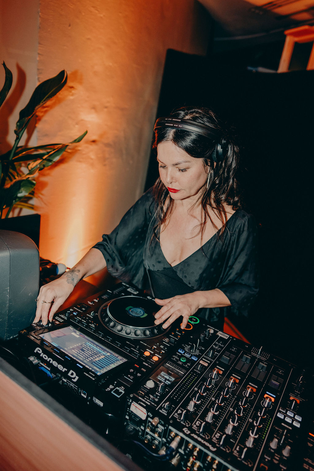 A woman wearing headphones and a black dress is DJing at a Pioneer DJ setup, adjusting controls on a mixer with warm lighting and a plant in the background.