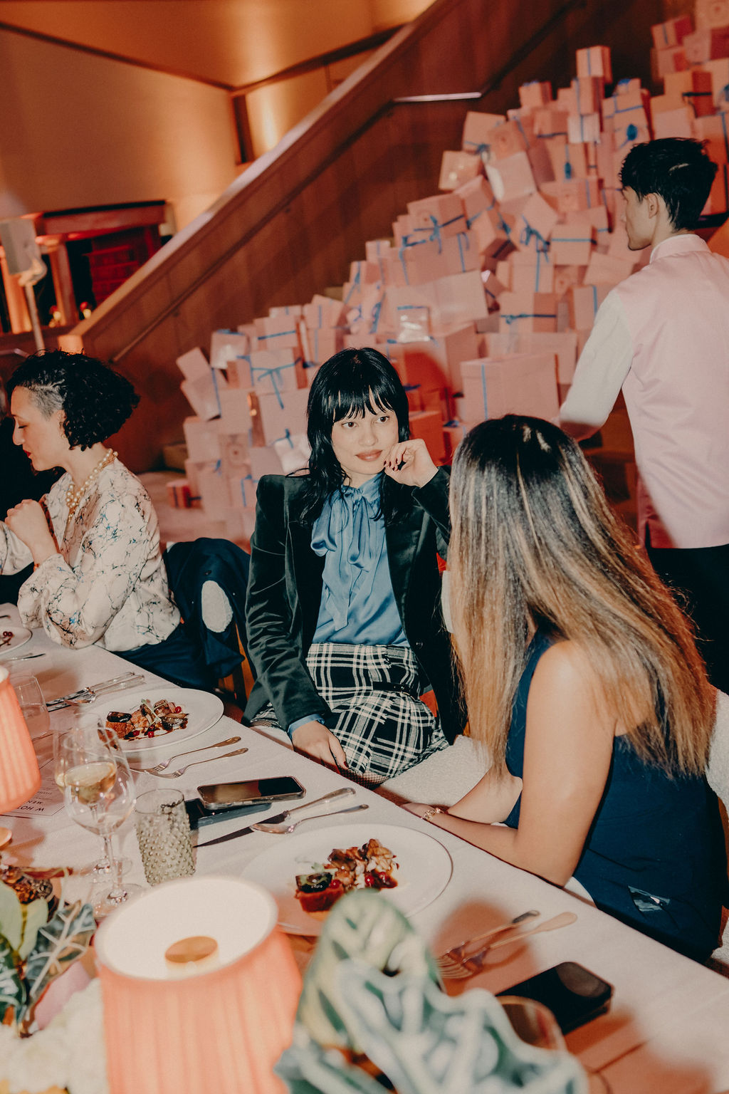 Three women are seated at an elegant dinner table, talking, with plates of food in front of them. Behind them, a staircase is covered with stacked white gift boxes tied with blue ribbons. A server stands nearby.