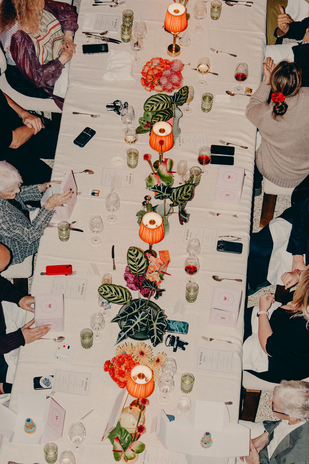 Overhead view of a long dinner table set with menus, glasses, phones, floral centerpieces, and red-shaded lamps. People seated on both sides are engaged in conversation and looking at menus.