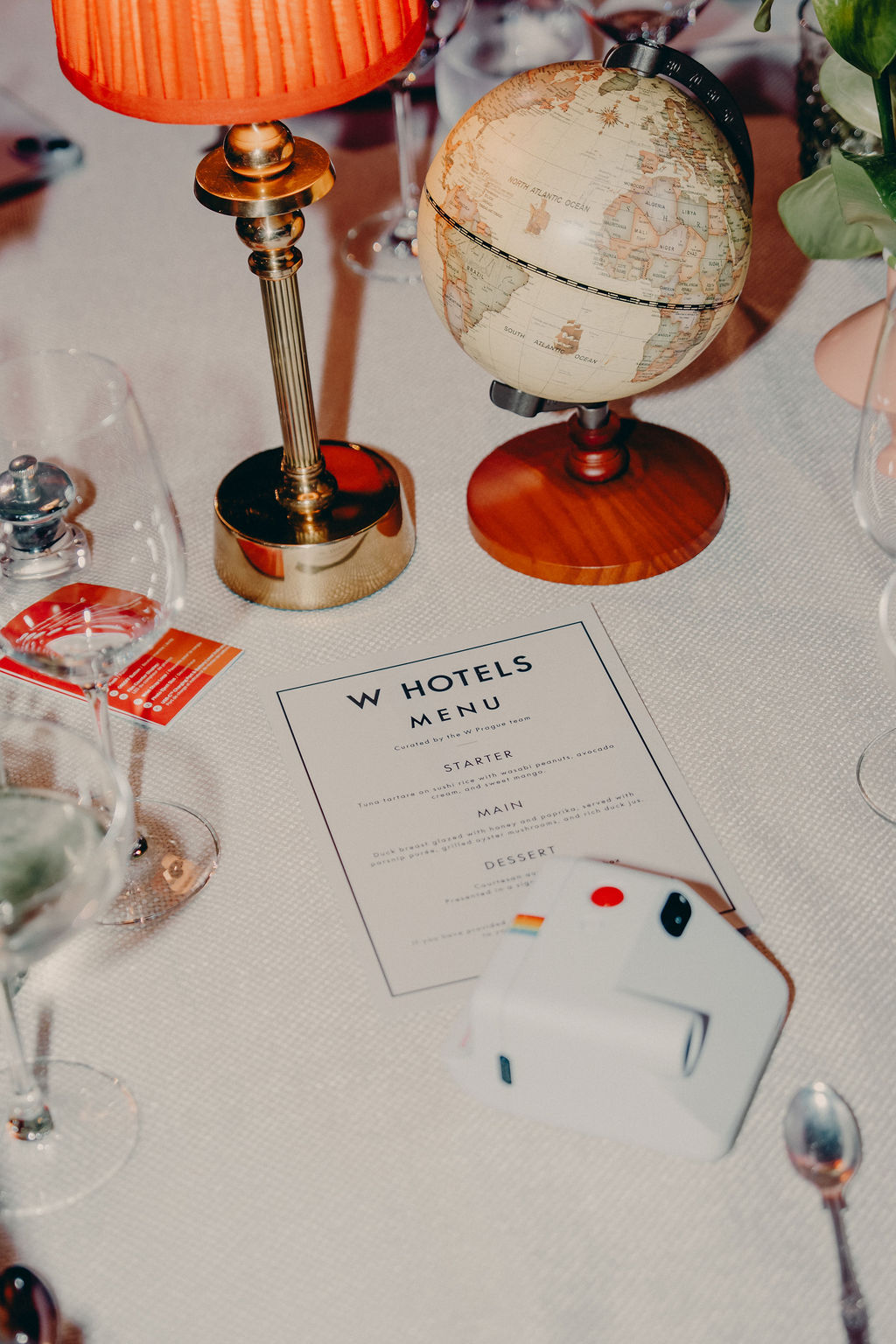 A table setting with wine glasses, a small globe, a vintage lamp, a W Hotels menu, and a white instant camera on a white tablecloth.