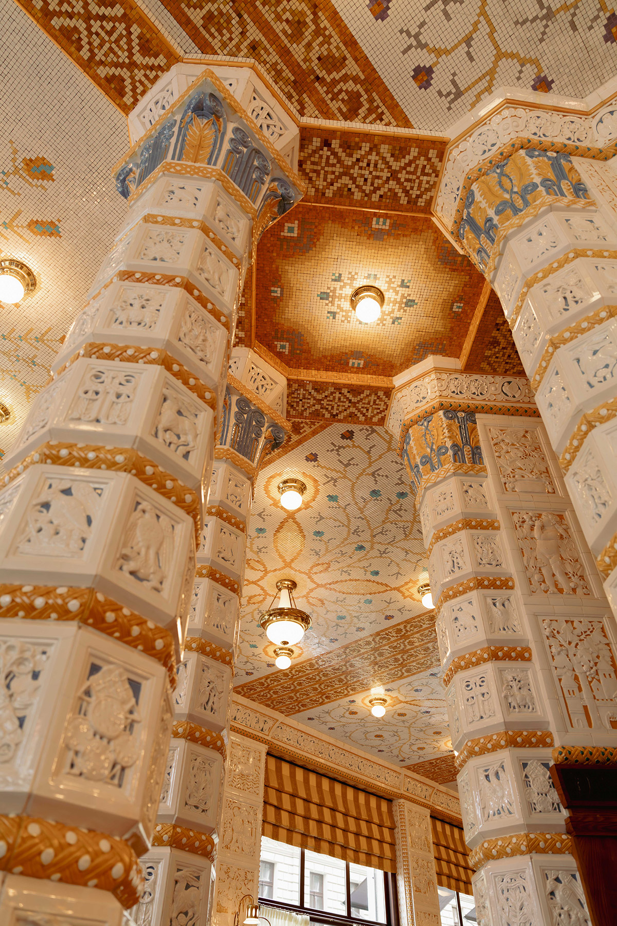 Ornate interior with tall, white columns decorated with gold and blue accents, and an intricately patterned mosaic ceiling with floral motifs and hanging lights. A large window is visible in the background.