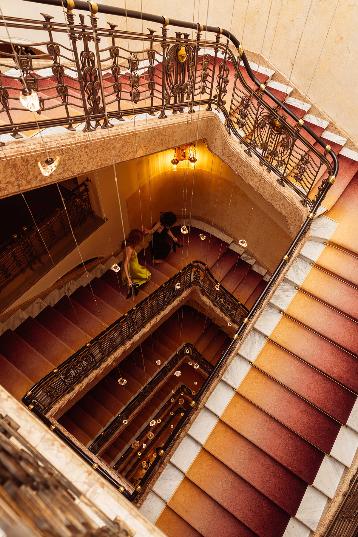 View from above of a spiral staircase with red and white steps, ornate railings, and hanging lights. Two people are seen sitting together on a lower landing, partially visible. The lighting creates a warm, elegant atmosphere.