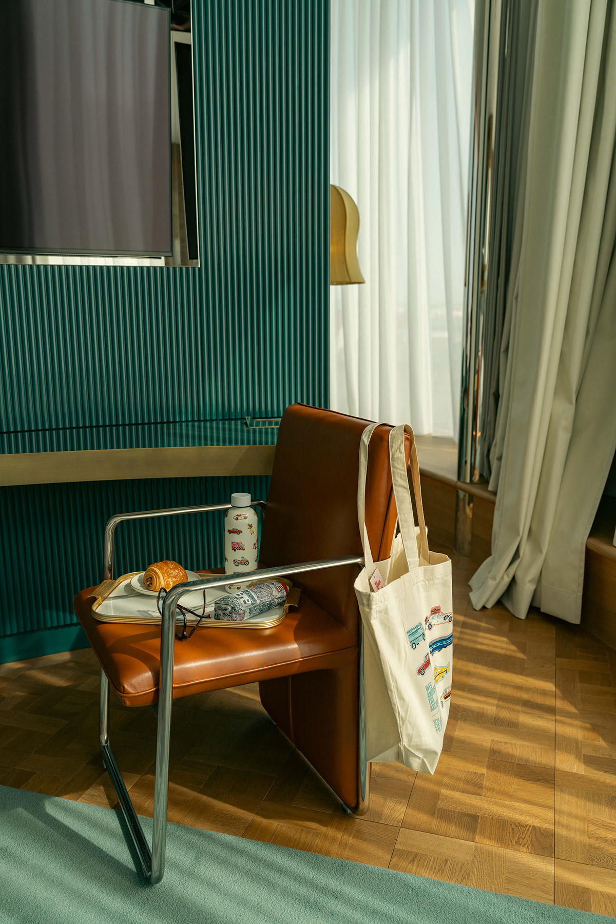 A modern brown leather chair with a metal frame holds a plate with a croissant, a can, and a bag. A cream tote bag with colorful graphics hangs on the armrest. Sunlight filters through nearby white curtains.