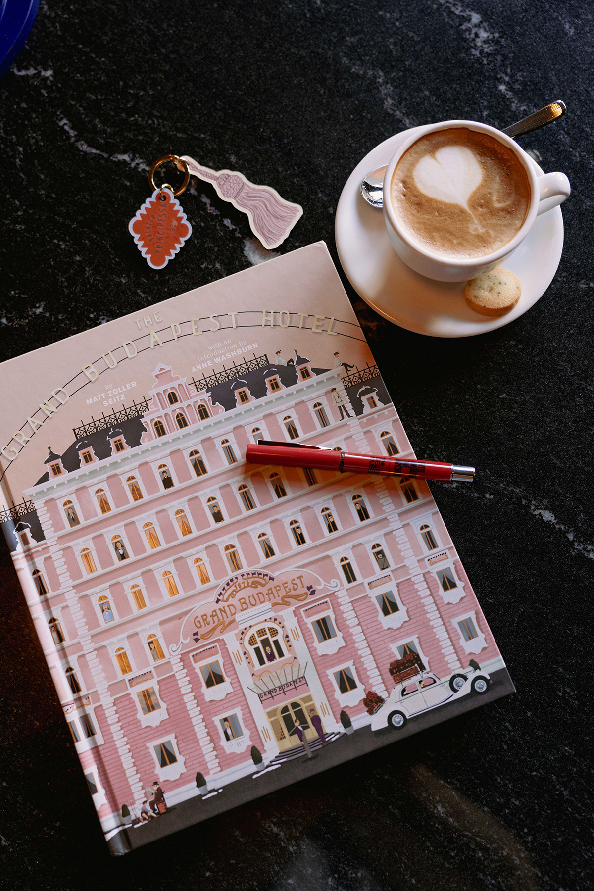 A notebook with an illustrated cover of The Grand Budapest Hotel, a red pen, a keychain with hotel-themed charms, and a cappuccino with a heart-shaped froth and a small cookie on a saucer, all on a dark surface.