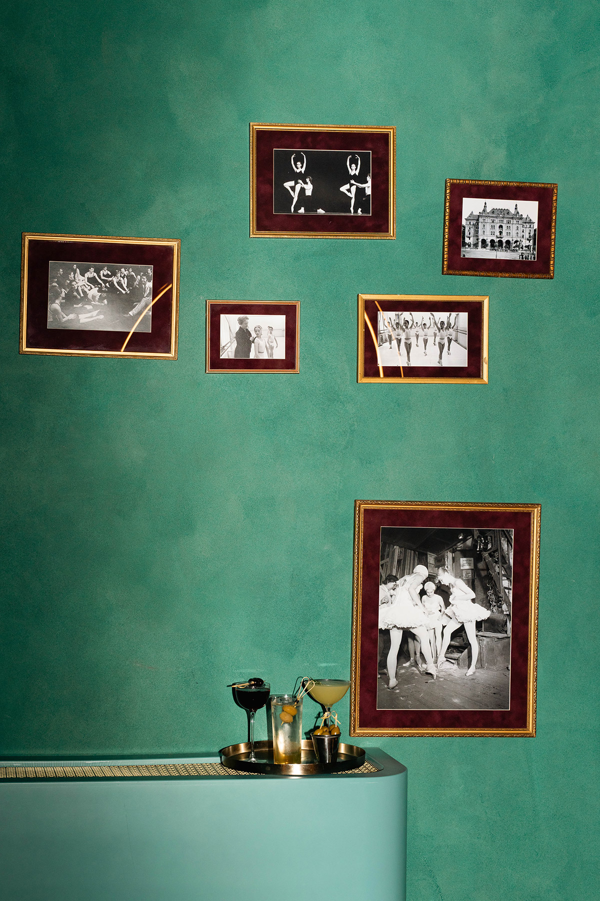 A teal wall displays six framed black-and-white photos of dancers and a theater. Below, a counter holds a tray with glassware and two drinks.