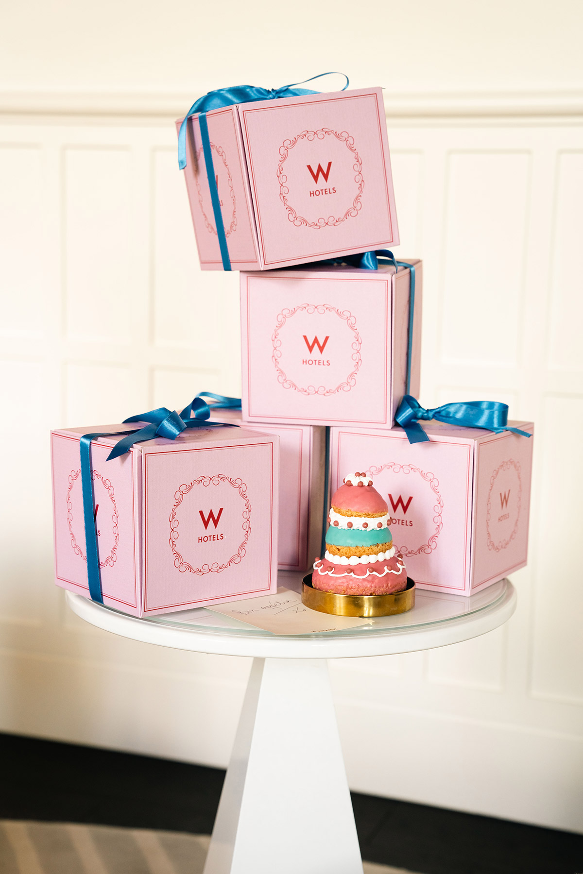 Four pink gift boxes with blue ribbons, branded with the “W Hotels” logo, are stacked on a round white table next to a small, colorful, tiered decorative cake. The background features white paneling.