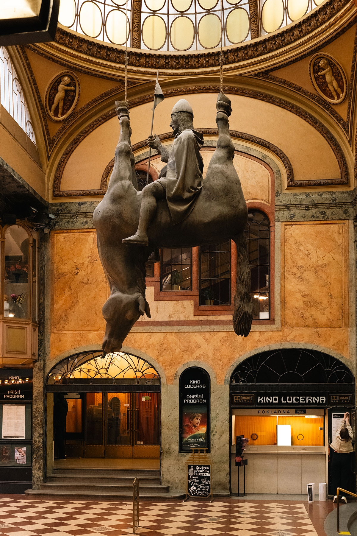 A large statue of a man in armor riding an upside-down horse hangs from the ceiling in an ornate, historic hall with arched windows, marble walls, and checkered floors.