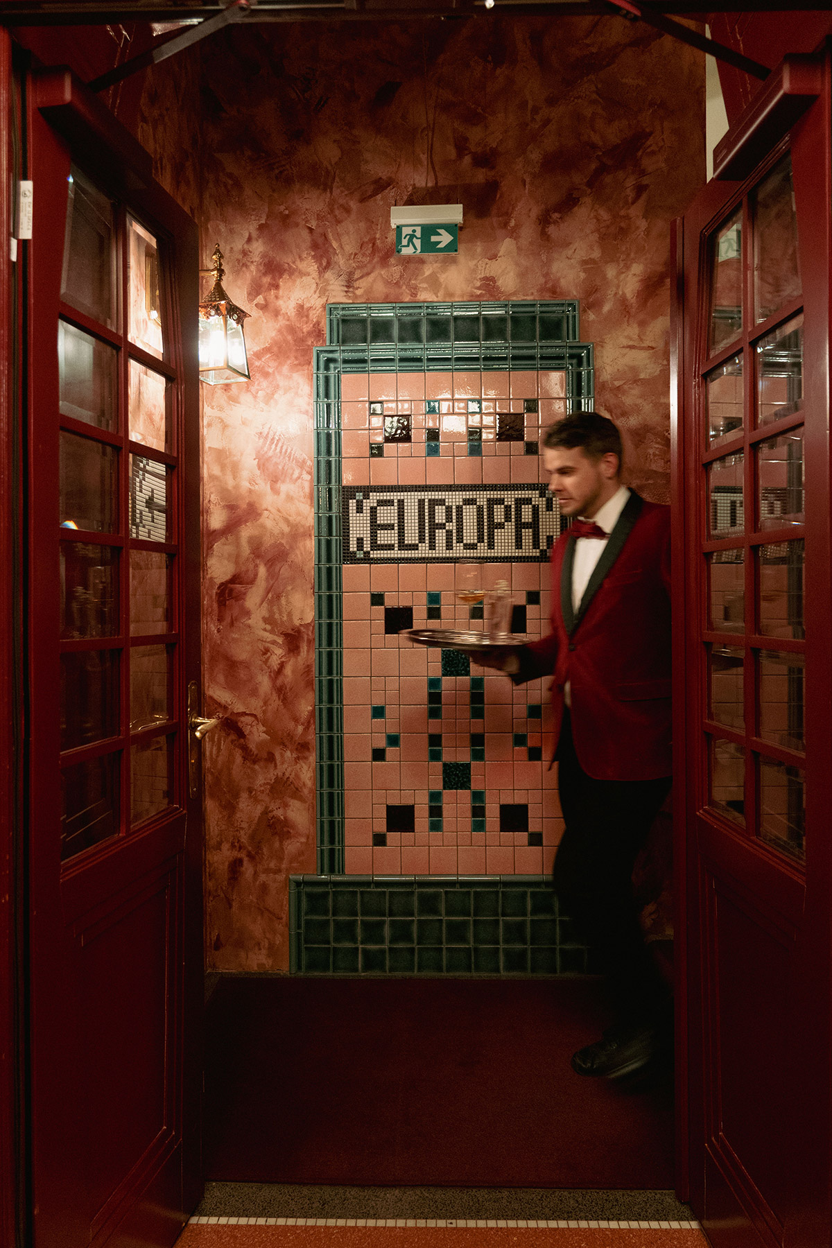 A man in a red jacket and bow tie walks through a doorway with glass panels, toward a wall with pink tiles and a green-tiled sign that reads 