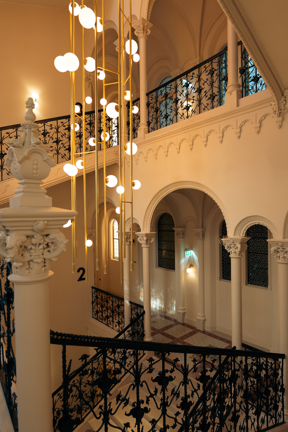A grand staircase with ornate black railings, arched doorways, and decorative columns. Modern hanging lights with round bulbs contrast with the classical architecture and cream-colored walls.