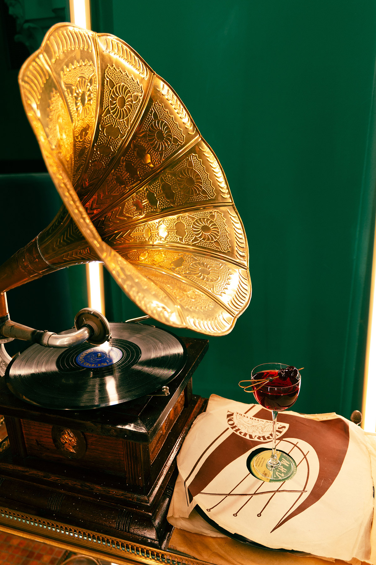 A vintage gold gramophone plays a vinyl record on a wooden table next to a cocktail glass filled with a dark red drink, garnished with a cherry, and a vintage record sleeve against a green background.