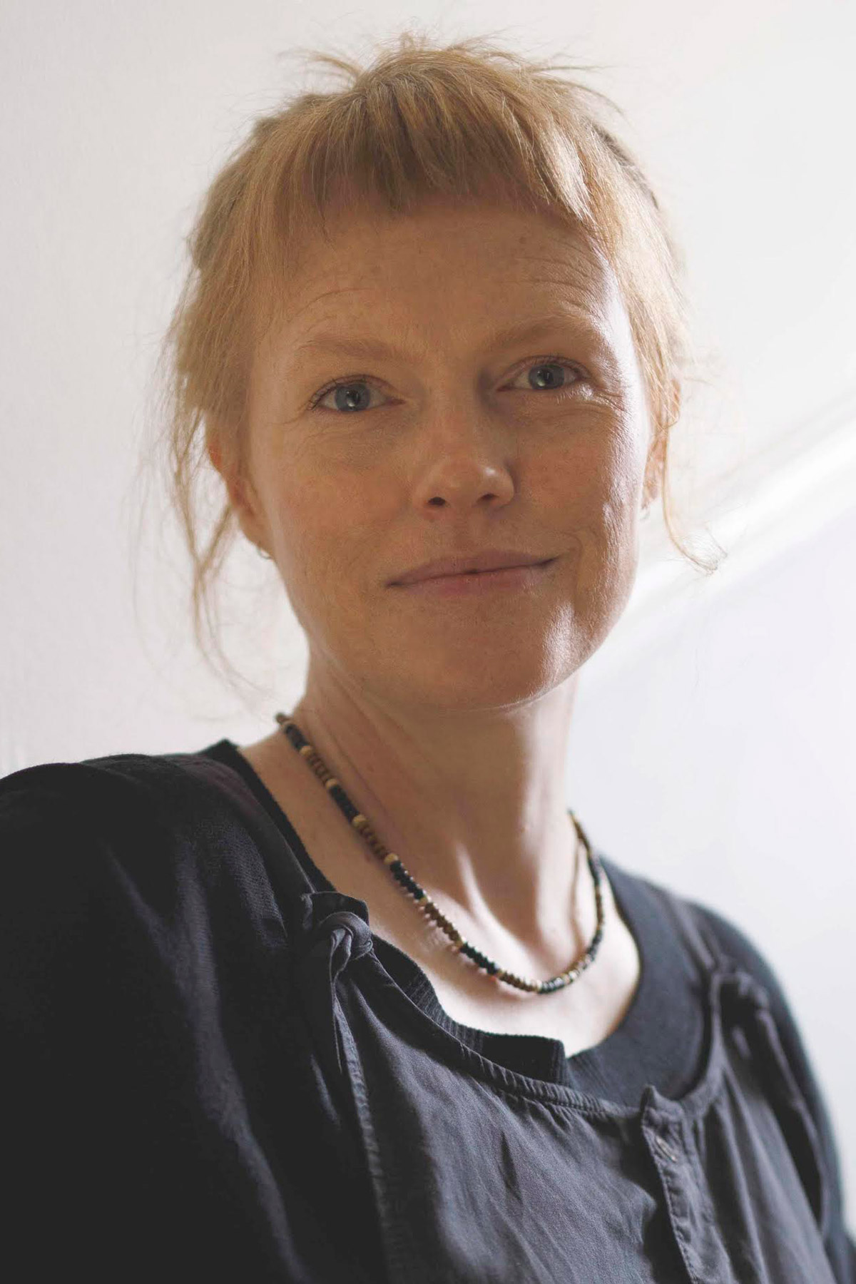 A woman with light skin and red hair, wearing a black top and beaded necklace, stands against a light background, softly smiling at the camera.