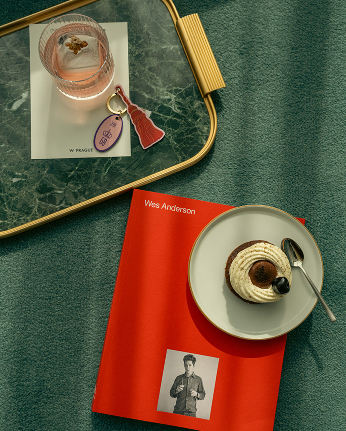 A red Wes Anderson book sits on a green surface with a plate holding a dessert and spoon on top. Nearby, a tray holds a glass of pink drink, a hotel keychain, and a card labeled 