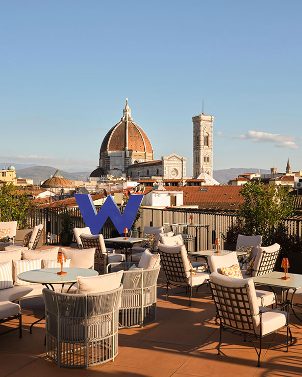 A rooftop terrace with cushioned chairs and tables overlooks Florence’s skyline, featuring the iconic dome of the Florence Cathedral and the bell tower. A large blue 