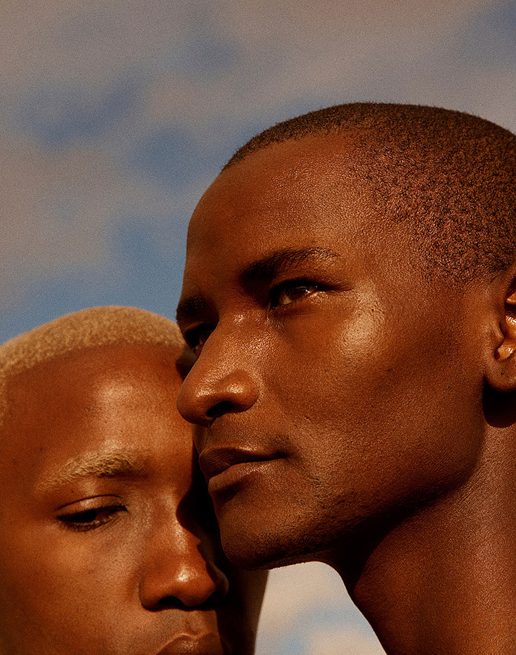 Two people stand close together under a blue sky; one with light blond hair has eyes closed, while the other with short dark hair looks thoughtfully into the distance. Warm sunlight highlights their skin.